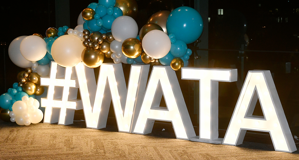 Illuminated “#WATA” sign with teal, white, and gold balloon decorations at an indoor event.