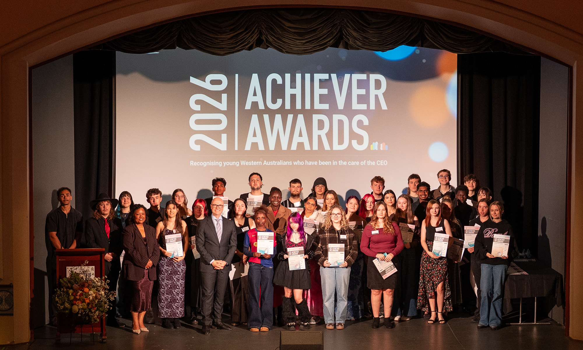 a large group of young people on stage holding their Achiever Awards with "2026 Achiever Awards" signage in the background