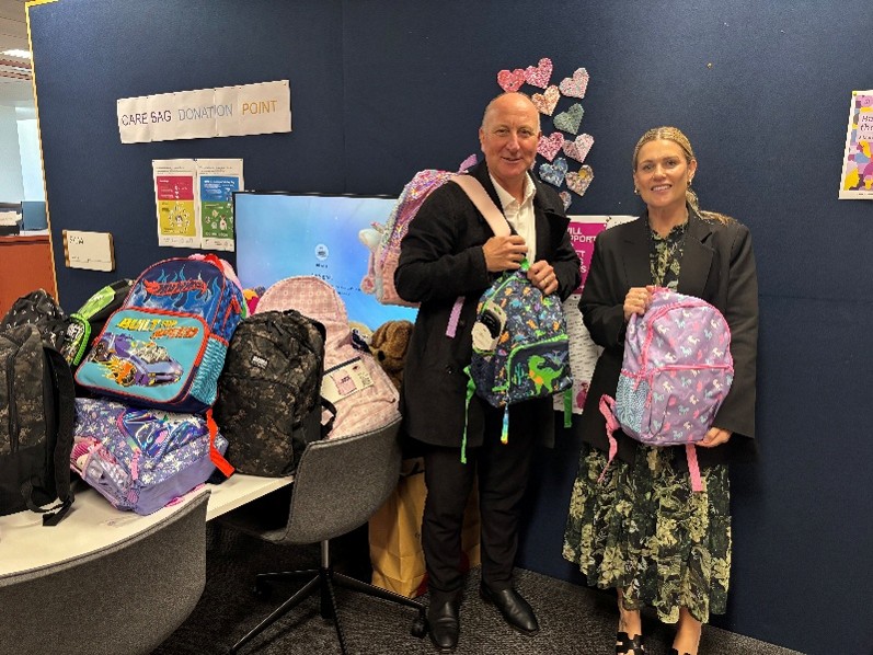 Two adults in an indoor setting surrounded by colourful bags filled with basic necessities 