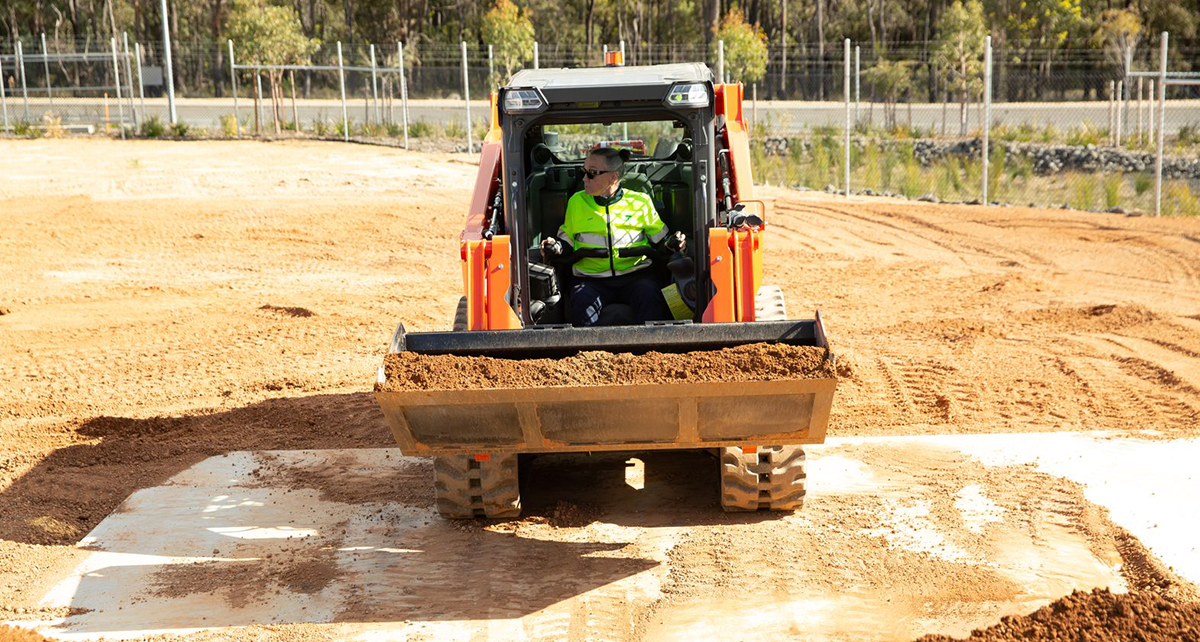 Person operating a skid steer loader carrying soil across a dirt training site.