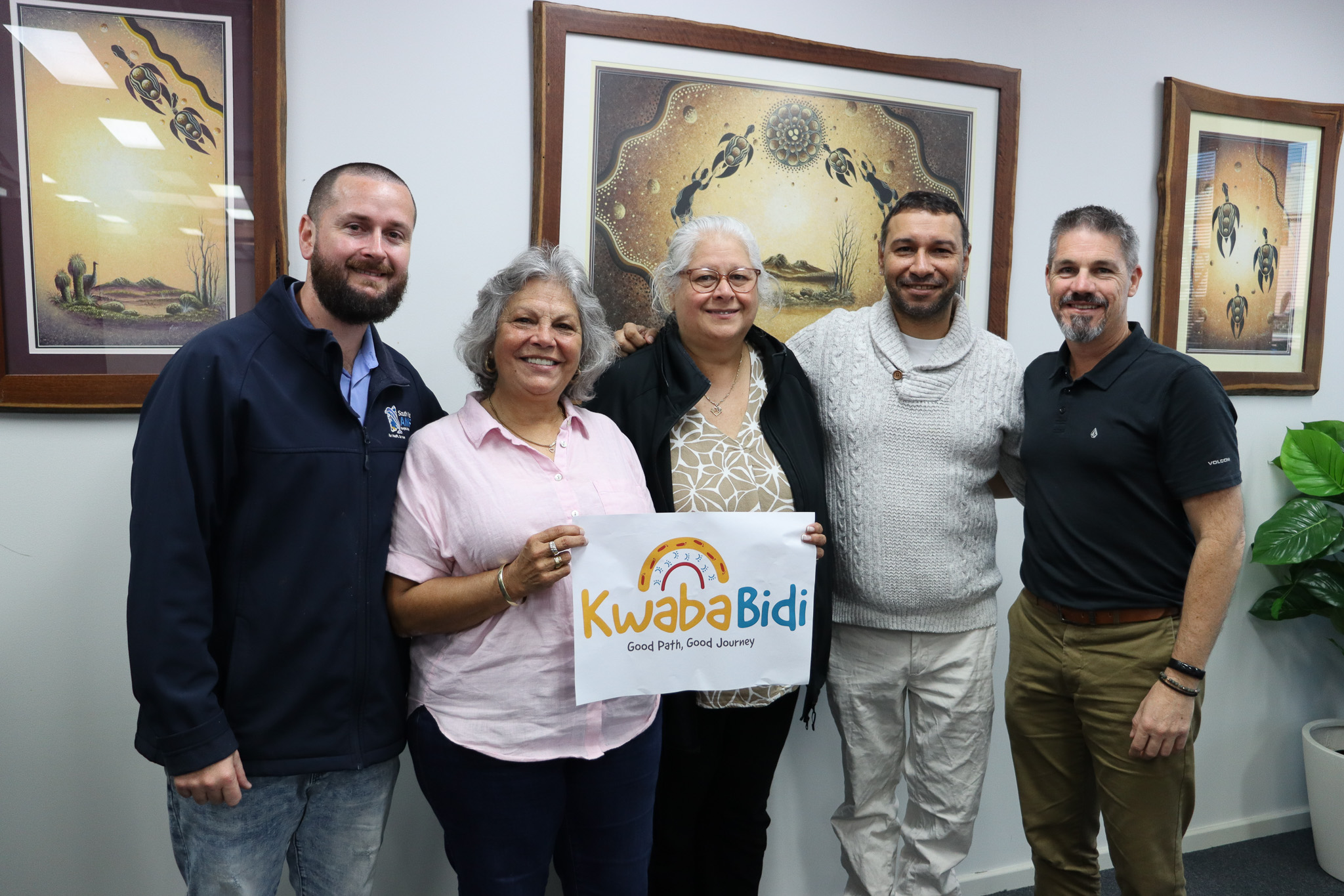 Five people stand together in an office beneath three framed artworks. Two people in the centre hold a sign reading “Kwaba Bidi – Good Path, Good Journey.” The group is posed for a photo against a white wall with Indigenous-style artwork.