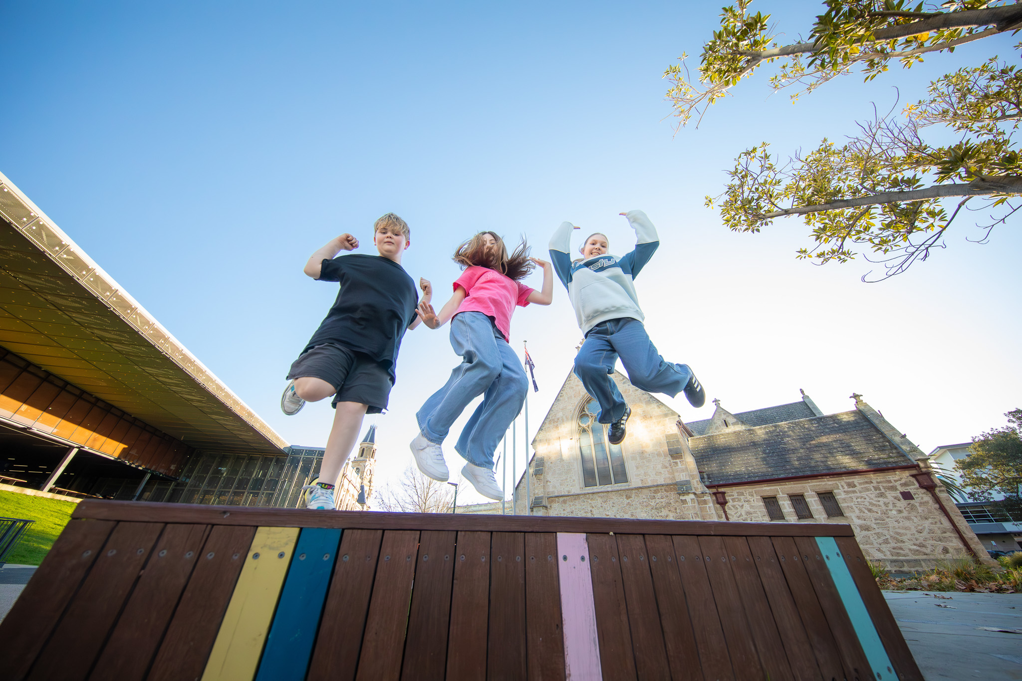 Three children jump off a colourful wooden bench in an outdoor plaza, with a historic stone building, modern architecture, and clear blue sky in the background.