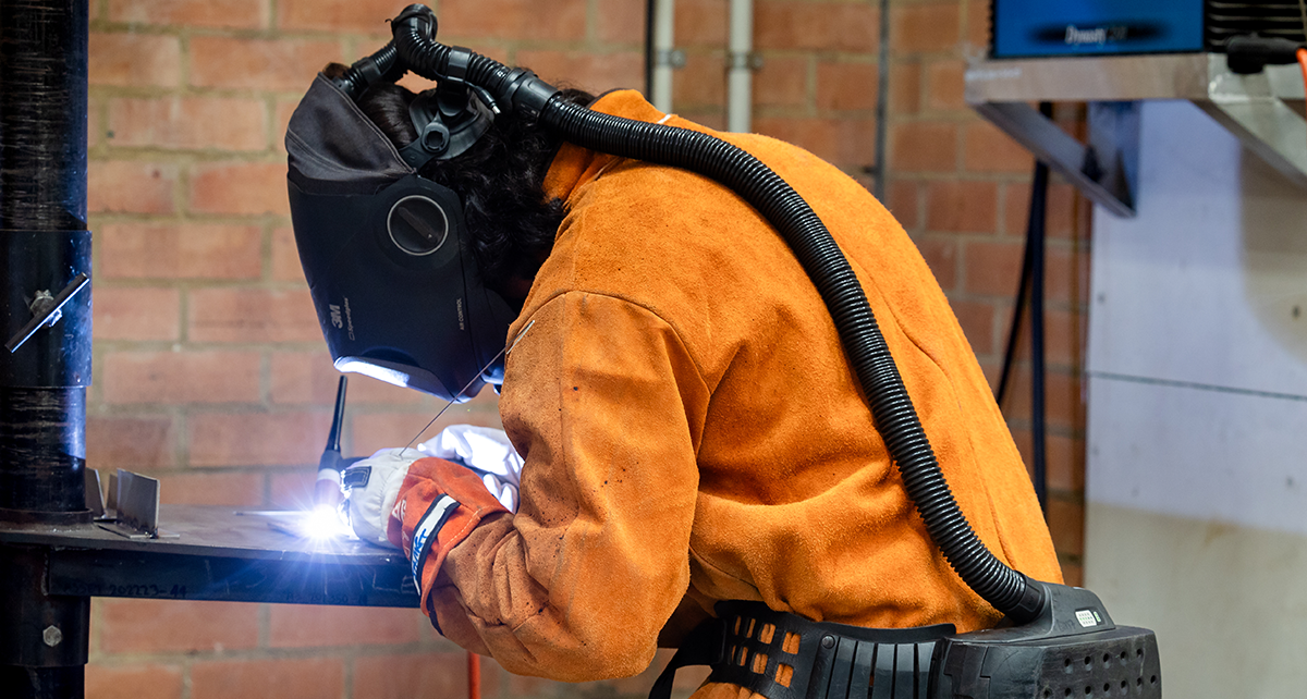 Student welding metal in a workshop while wearing protective safety gear.