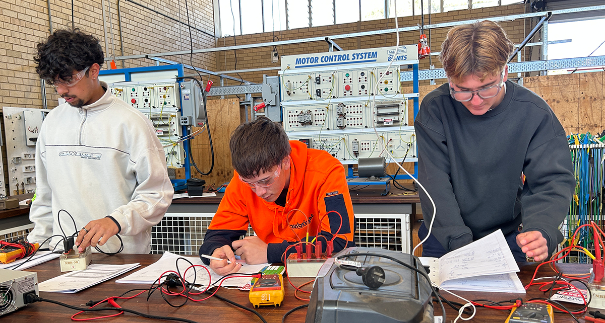 Three students working on electrical training exercises in a workshop, surrounded by wiring, tools and testing equipment.