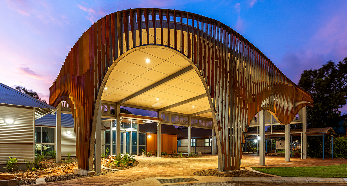 Entrance to a North Regional TAFE campus featuring a distinctive curved steel canopy, illuminated at dusk with landscaped gardens and paved walkways.
