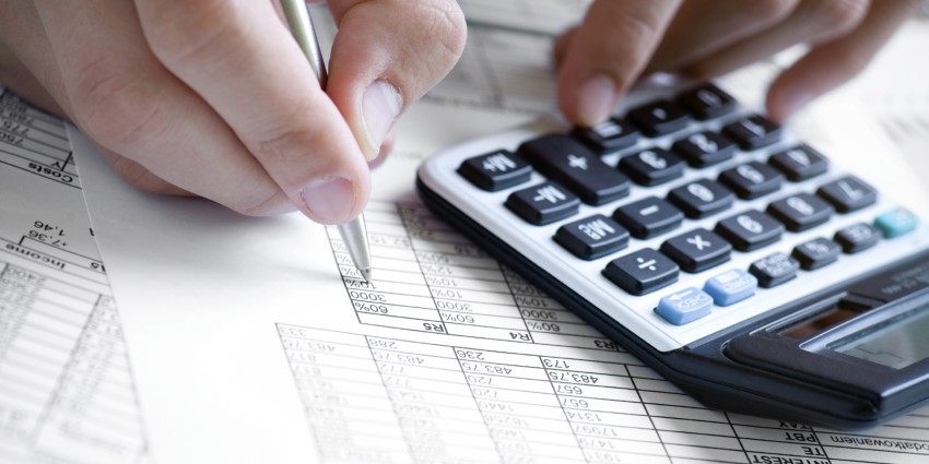 Hand holding a pen checking a balance sheet on a calculator.