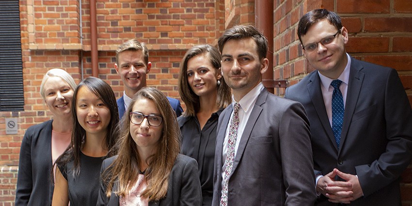 Seven Finance graduate employees standing in front of a brick wall.