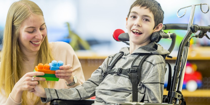 Young boy in wheelchair smiling. He is with a young female carer who is holding colourful building blocks.