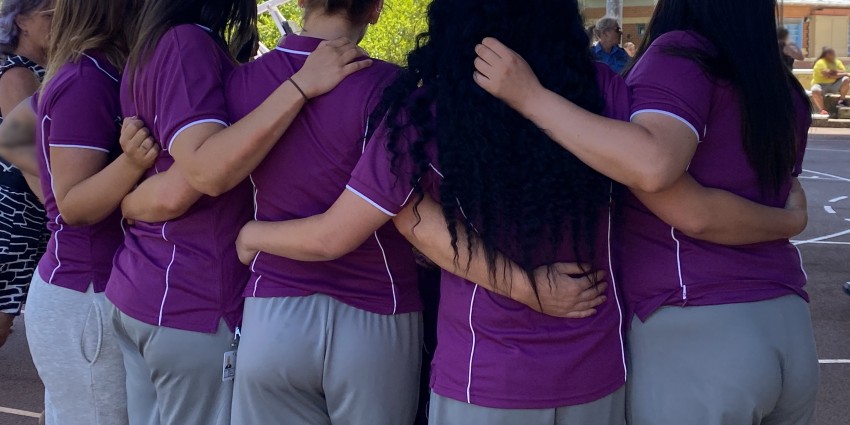 A group of women wearing purple tops and grey shorts are standing in a group with their arms around each other. The women have dark hair and are standing outside on a sunny day with blue sky.