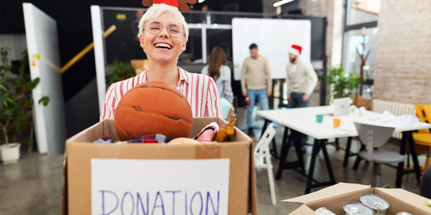 A volunteer wearing a Christmas head piece, smiling. She is standing in front of a box labelled donation, which is full of toys.
