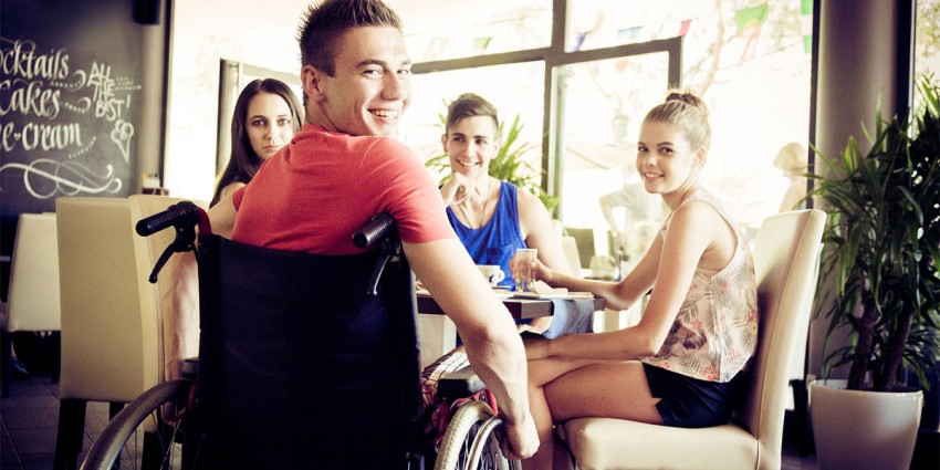 A young man in a wheelchair, smiling with his friends at a cafe.