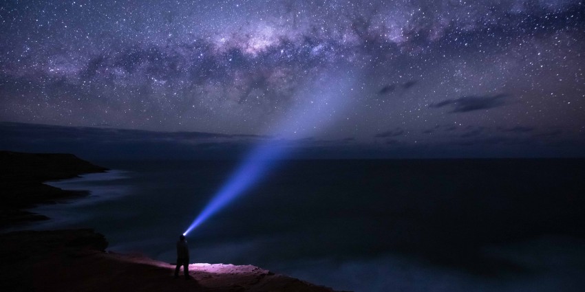 Person with a headlamp standing on the edge of a cliff looking out at the stars in the night sky