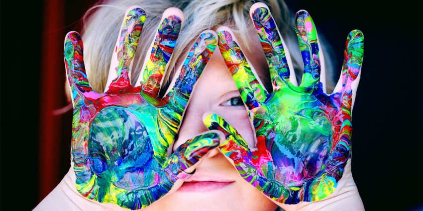 Young child holding hands to her face. Her hands are covered in colourful paint.