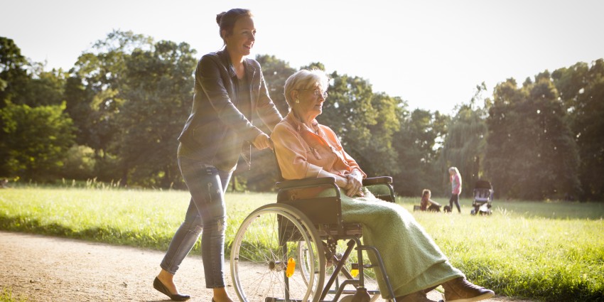 A young woman pushing an older woman in a wheelchair. They are in a park, enjoying the sunshine.