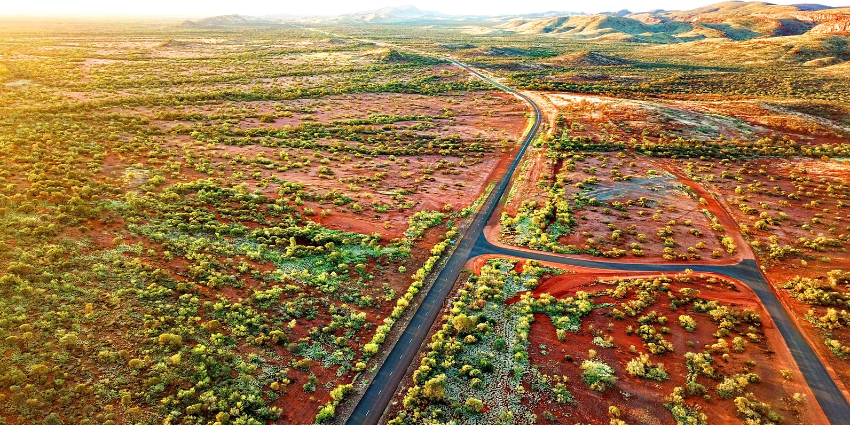 Image taken from above of a road running through wide flat red earth covered with scrubby green bush 