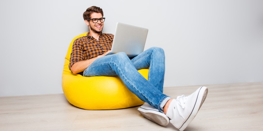 Happy young man sitting in yellow pouf and using laptop