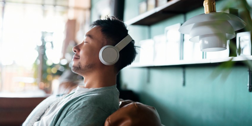 Young Asian man with eyes closed, enjoying music over headphones while relaxing on the sofa at home