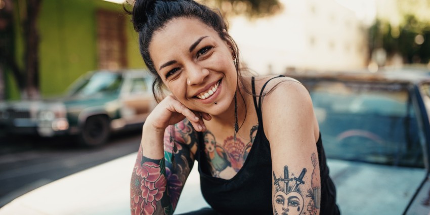Tattooed young adult woman smiling outdoors and sitting on a car hood 
