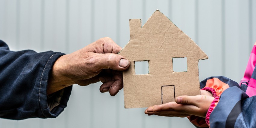 A man's hand holds a paper house cut out of cardboard. Nearby, children's hands hold the house from below.