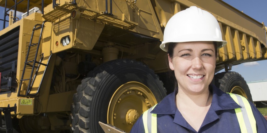 Female mining employee wearing high visibility vest, protective hard hat and holding clip board, smiling in front of a large mining truck