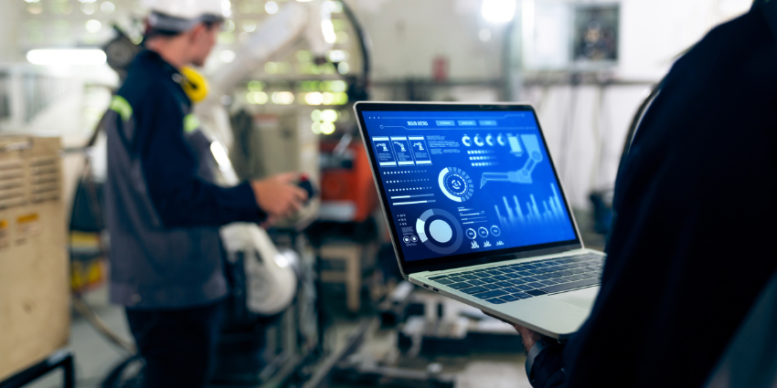 Close-up of a worker holding a laptop with graphs and data visible on the screen. In the background is a factory worker in a helmet