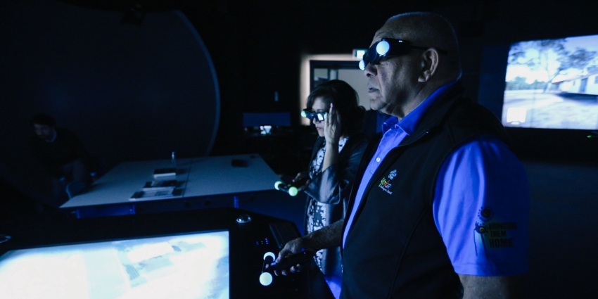 Two individuals using virtual reality equipment. They are in a dark room with light from a screen illuminating their faces. 