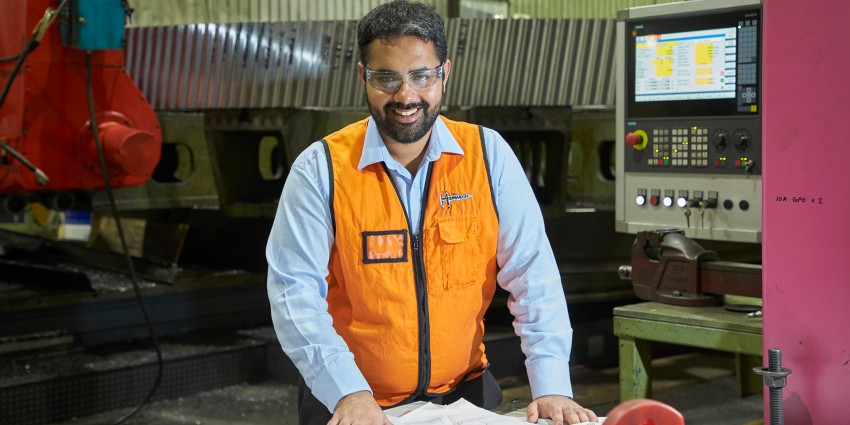 An engineer wearing an orange vest and protective eyewear, in a warehouse, smiling at the camera.