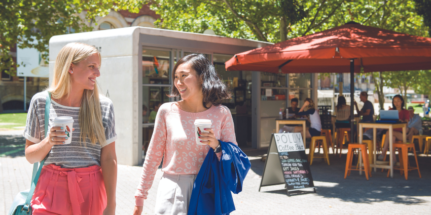 two girls with takeaway coffee cups looking at each other smiling with a cafe in the background