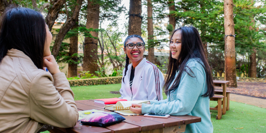 three girls sitting at table smiling with notebooks in front of them and pine trees in the background