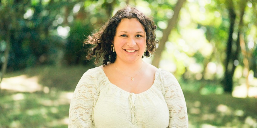 A person with curly hair, wearing a light lace blouse and necklace, smiles while standing outdoors with greenery in the background.