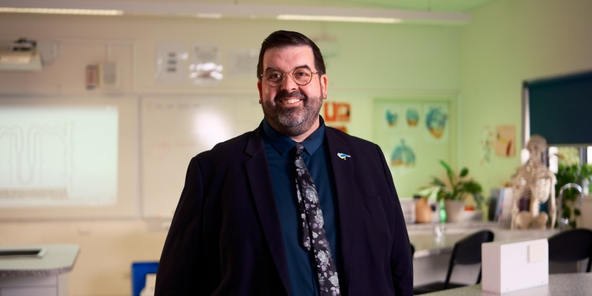 A man in a suit and tie stands confidently in a classroom, surrounded by desks and educational materials.