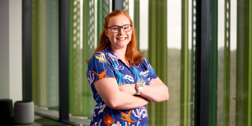 A woman in glasses and a floral shirt stands near a window, framed by soft light that enhances her cheerful appearance.
