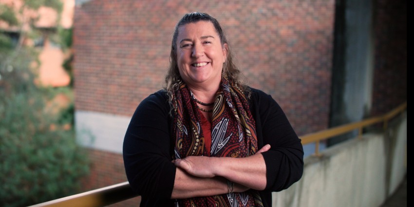 A person with long hair smiles while standing on a balcony with arms crossed, against a backdrop of brick buildings and greenery