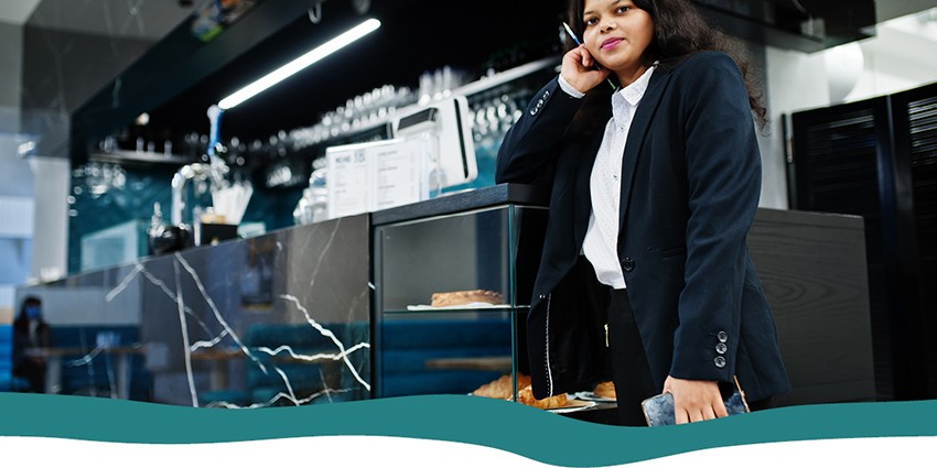A lady in a black suit leaning against a cafe/restaurant counter with a notebook and pen in hand