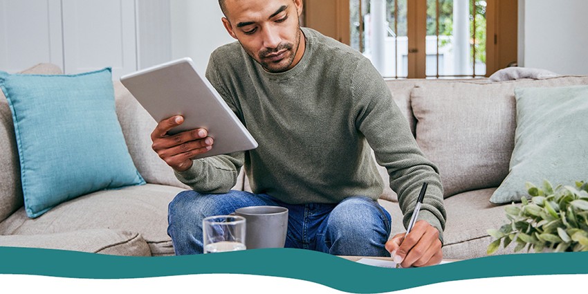 A man sitting on his couch with tablet in hand filling out a form on paper