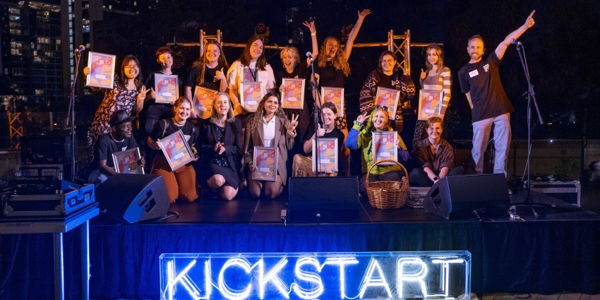Group photo of 16 young adults on stage with the word Kickstart lit up in front of the stage
