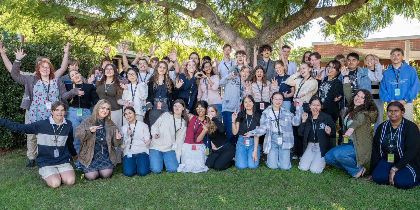 A group photo of a big group of young people aged 15 to 25, pictured in an outdoor setting on green grass with a tree in the background 