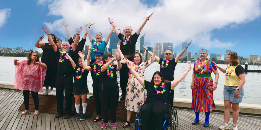 A cheerful group of people, including one person in a wheelchair, pose with raised arms on a wooden deck beside a waterfront. They wear colourful leis, and a city skyline is visible in the distance beneath a blue sky with scattered clouds.