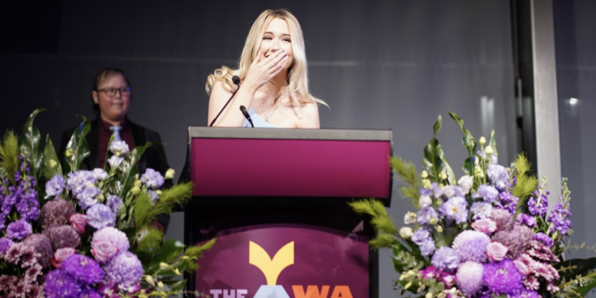 A young person at a podium with flowers, smiling and covering their mouth during the 2025 WA Youth Awards..