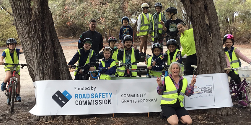 A group of children with bicycles amongst trees smiling and posing with a woman in the foreground in front of a banner that says "Funded by Road Safety Commission Community Grants Program"