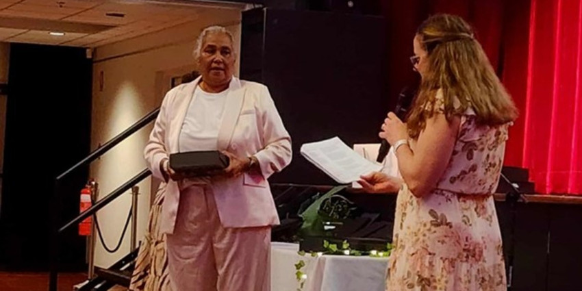 Two women are on a stage. One is receiving an award, the other is holding a microphone and reading a piece of paper.