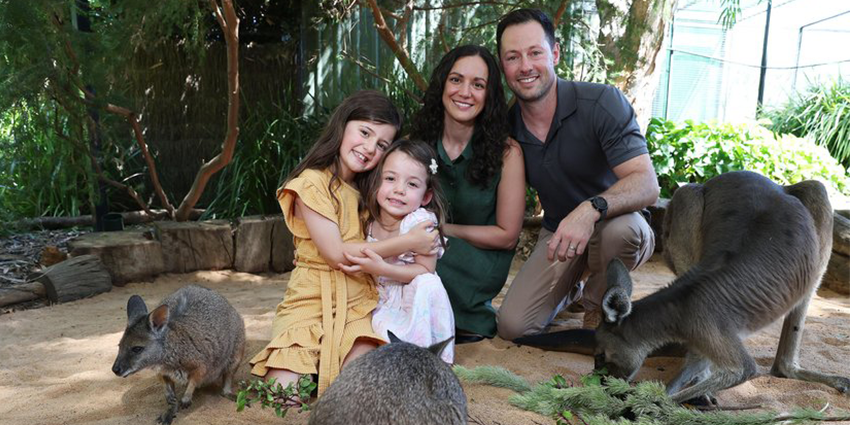 A man and women are with two young girls at a wildlife park. They are crouched down and smiling, surrounded by three kangaroos that are feeding on branches.