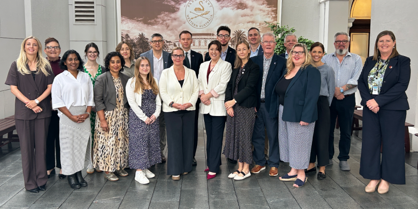 A group of people standing together in a courtyard, posing for a professional group photo in front of a building with columns and a large emblem.