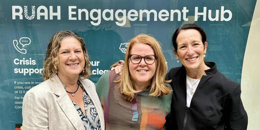 Three women smiling to camera with a banner behind them that reads "Welcome to the RUAH Engagement Hub"
