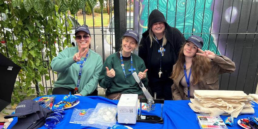 four young women, smiling, wearing lanyards, sitting at a table set up to provide information