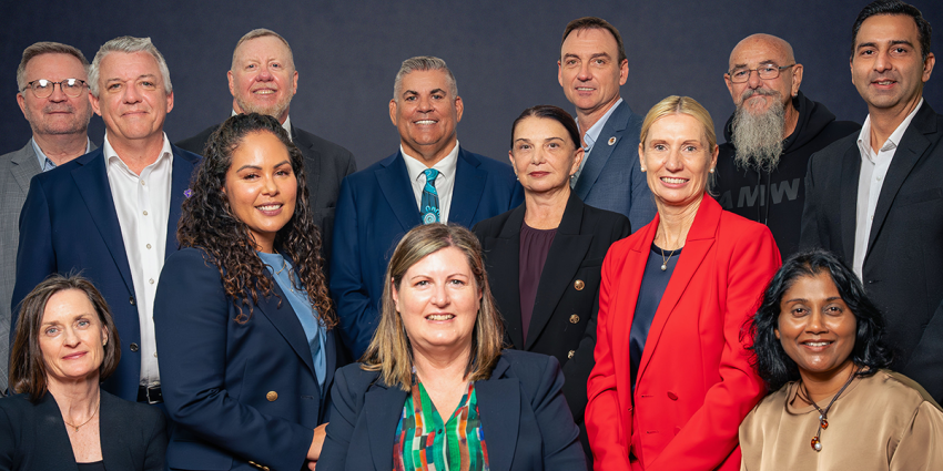 Group portrait of advisory board members standing and seated in two rows against a dark studio background, wearing professional attire and facing the camera.