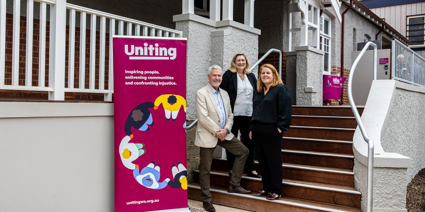 three adult people standing on the front steps of a character home, beside a colourful Uniting WA banner