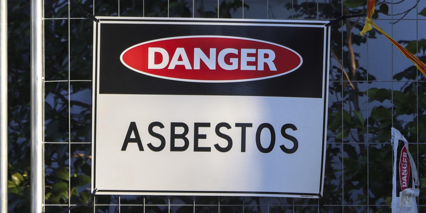 The image shows a close-up of a safety warning sign attached to a temporary wire fence outdoors which says Danger, Asbestos.