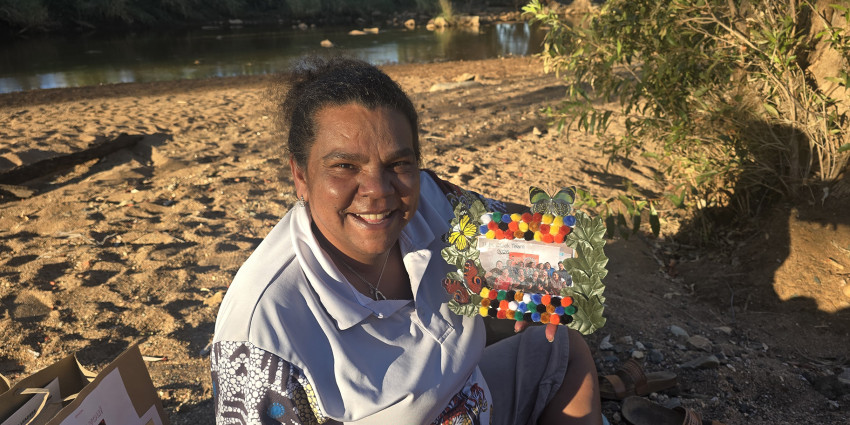 a woman smiling, holding flowers, wearing a polo shirt with Aboriginal artwork. She is sitting on the sand in a natural outback WA environment.