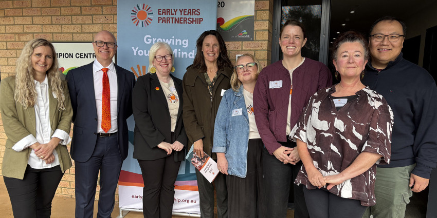 Group shot of seven neatly dressed adults in an outdoor setting, with an Early Years Partnership banner in the background